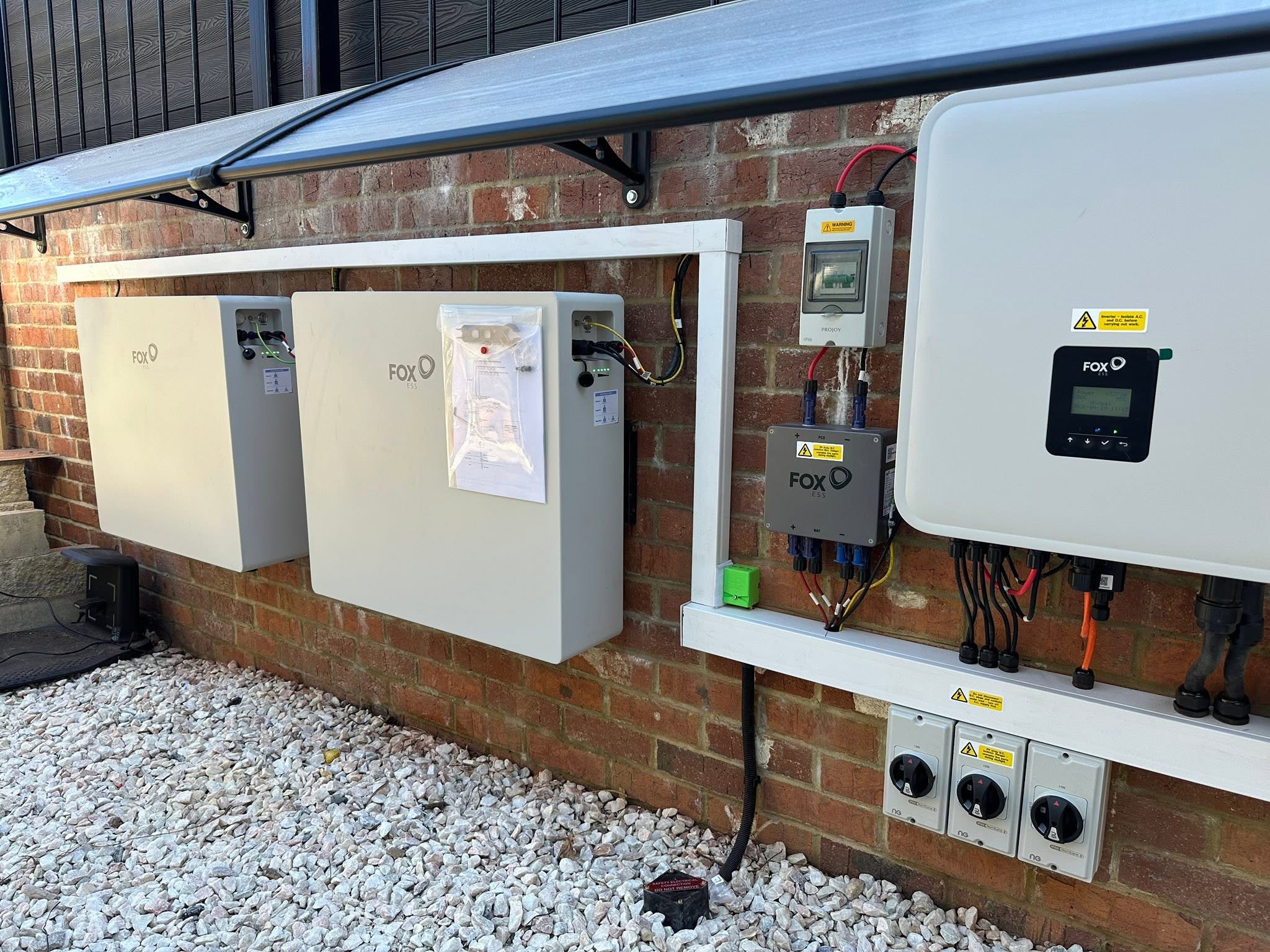 Two Fox ESS batteries and a hybrid inverter fitted on an outside wall under a canopy, with isolators and trunking