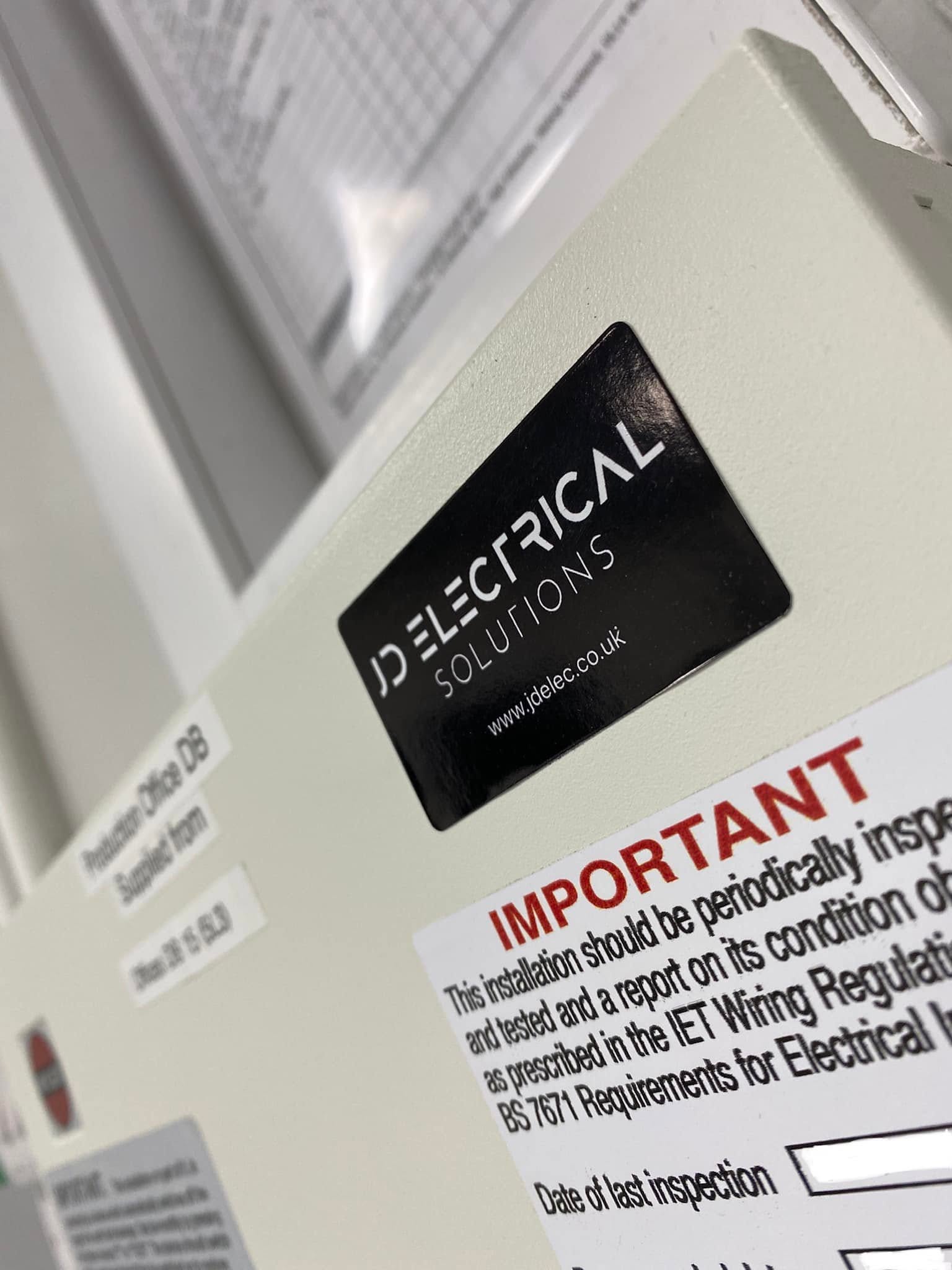A JD Electrical Solutions sticker beside the periodic inspection label on a distribution board